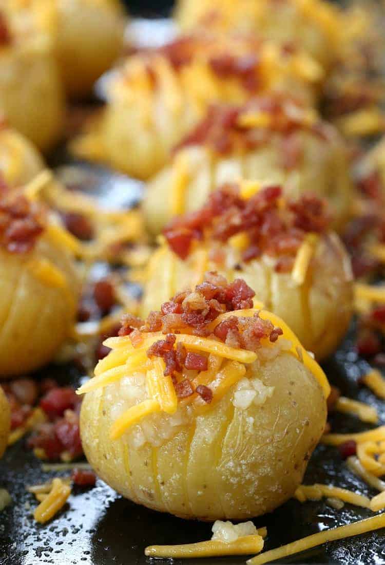 A tray of mini loaded hasselback potatoes, topped with garlic, shredded cheese, and bacon bits, ready for the oven