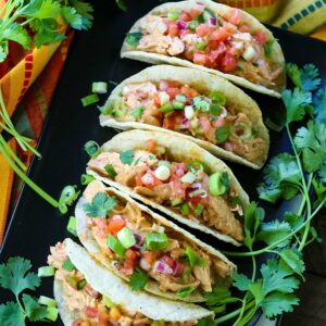 Creamy salsa chicken tacos with tomato and onion lined up on a plate surrounded by cilantro.