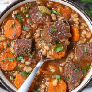 spoon in bowl of beef and barley soup with rolls on side