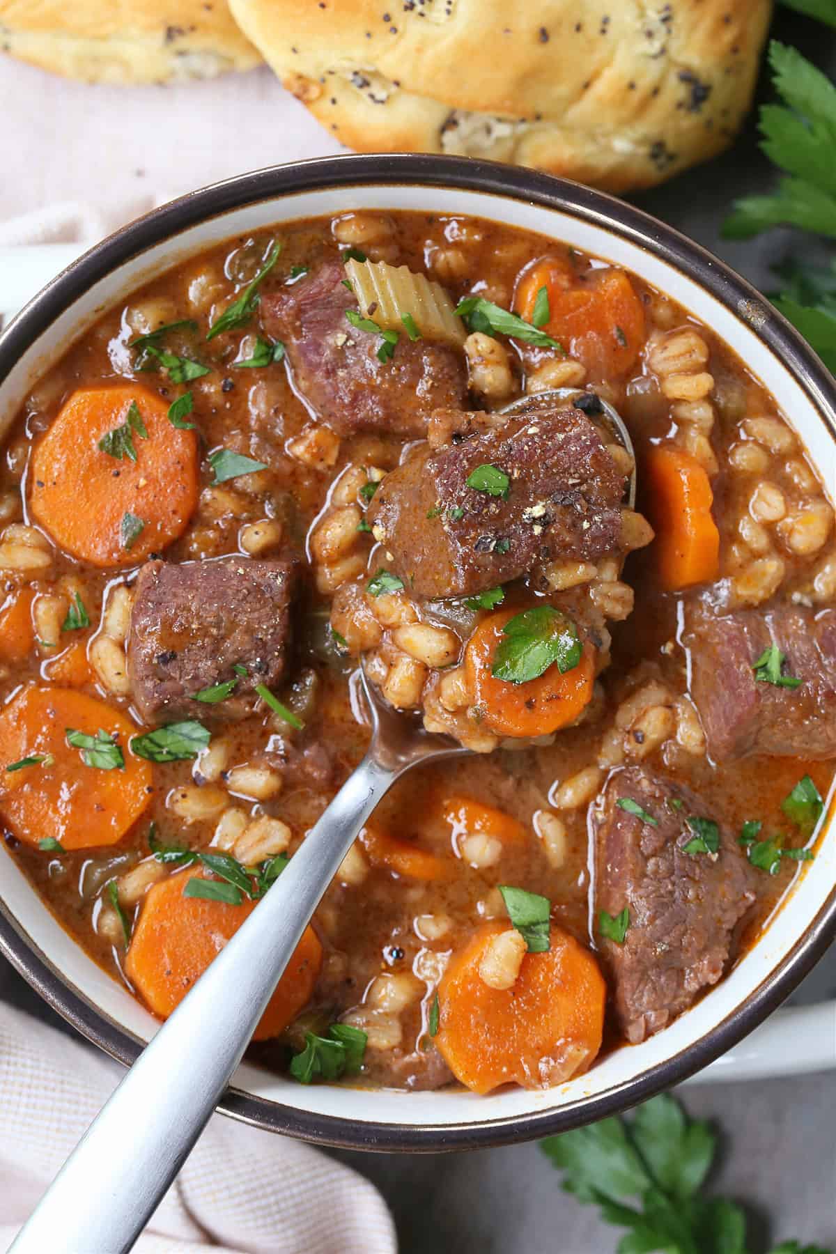 spoon in bowl of beef and barley soup with rolls on side