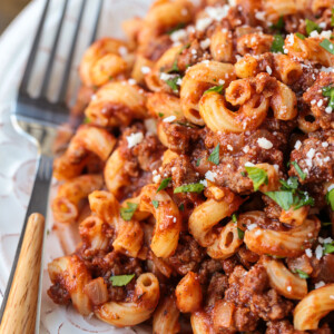 Close up of beefaroni on a plate, with parmesan and parsley on top, and a fork on the side
