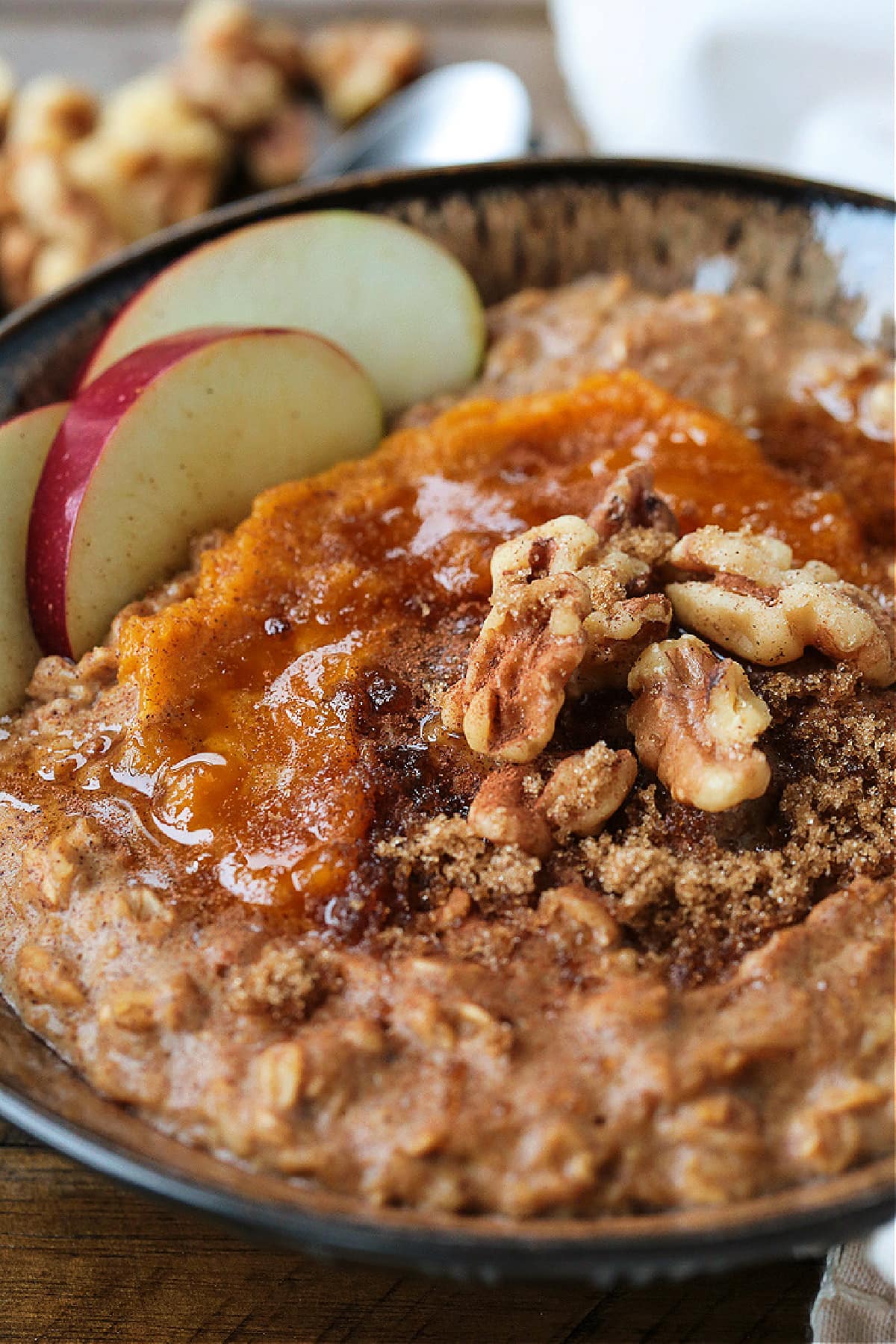 A bowlful of pumpkin oatmeal topped with apples, walnuts and pumpkin puree, with a spoon.